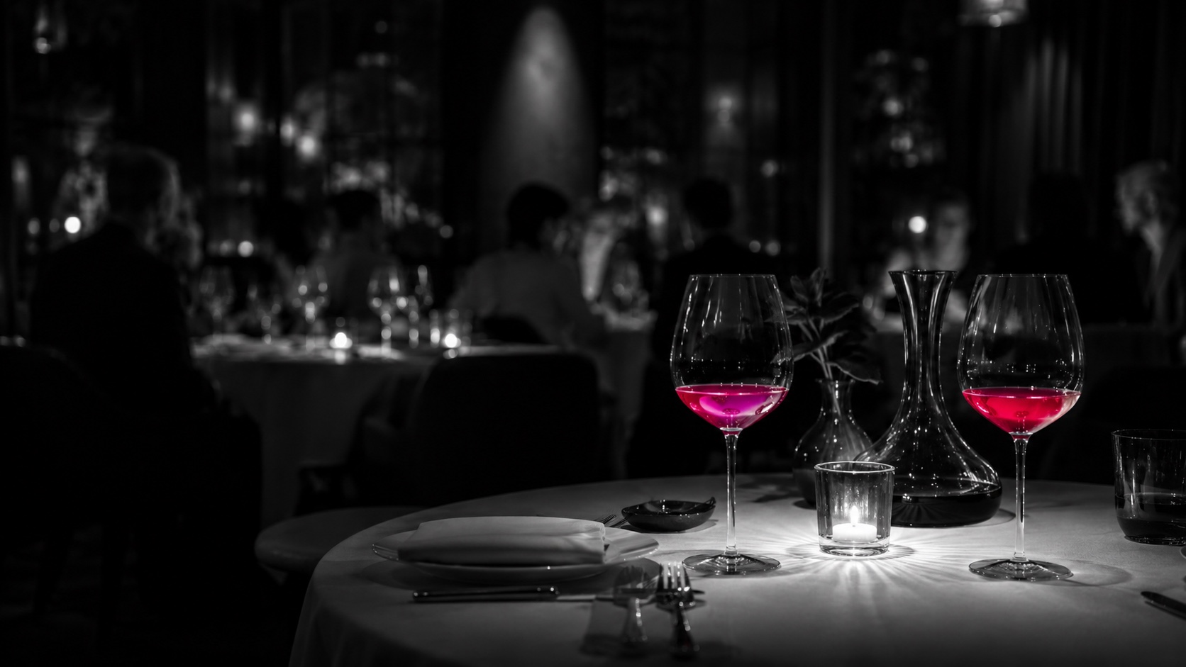 Sala de un restaurante en penumbra por la noche, en blanco y negro, las copas de vino brillan en magenta