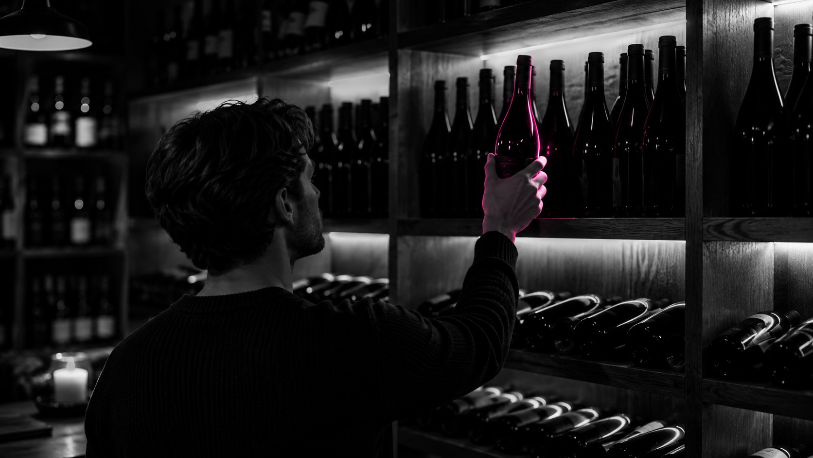 Mann greift im dunklen Weinladen nach einer magenta schimmernden Flasche, Schwarzweiß